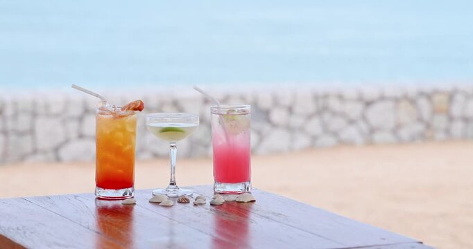 Tropical cocktails sitting on wooden table, sandy beach background with stone wall and ocean, summer vacation and luxury resort marketing