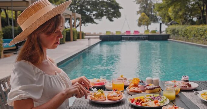 Woman wearing straw hat eating eggs benedict and fresh fruit, luxury tropical resort poolside with ocean view background, travel lifestyle, holiday dining, brunch marketing