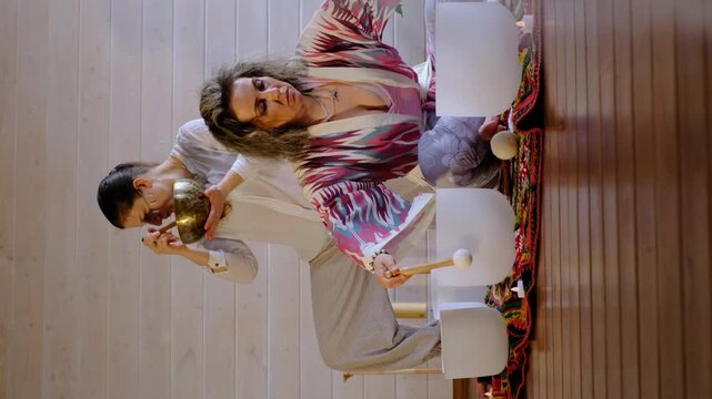 Eye level view of two women kneeling on a colorful rug as one guides the other in playing crystal bowls. Sound bath practice brings relaxation, stress relief, and calm focus to shared wellness.