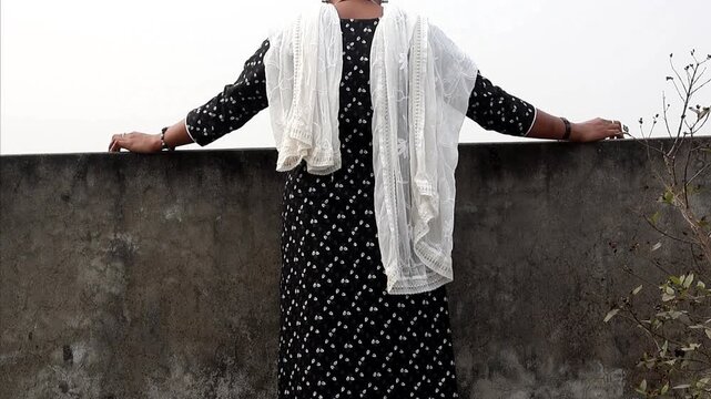 Back view of a young Bangladeshi woman in a black patterned salwar kameez and white dupatta, leaning on a rooftop ledge and looking at the city landscape. Concept of solitude, peace, and lifestyle
