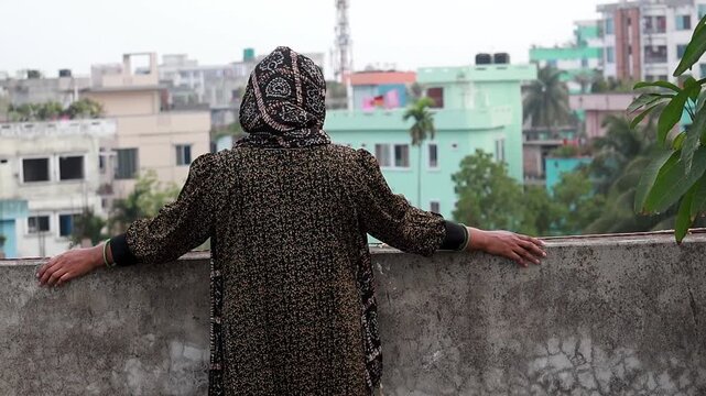 Back view of a woman with a covered head standing on a rooftop looking at the city. This shot highlights urban lifestyle, personal reflection, and peaceful moments in the city.