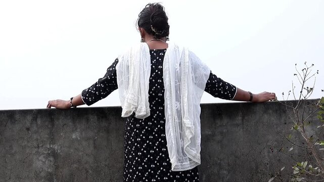 Back view of a young Bangladeshi woman wearing a black patterned salwar kameez and white dupatta, leaning on a rooftop ledge looking at the urban city view. Solitude and peaceful lifestyle concept