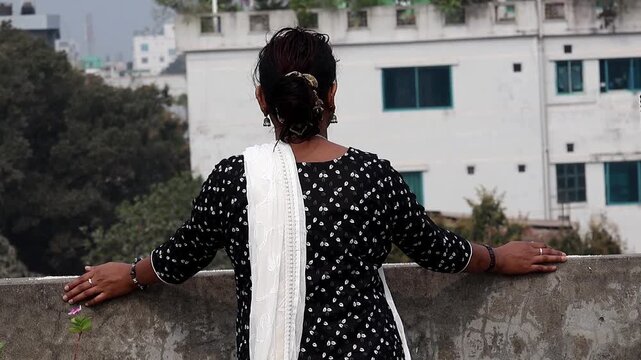 Back view of a Bangladeshi woman in a black patterned salwar kameez and white dupatta, leaning on a rooftop ledge and looking at the urban city landscape. Concept of solitude, peace, and lifestyle