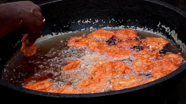 Hand dropping raw lentil batter into hot boiling oil to fry crispy onion fritters or pakoras. This scene represents authentic South Asian street food culture and traditional deep frying methods.