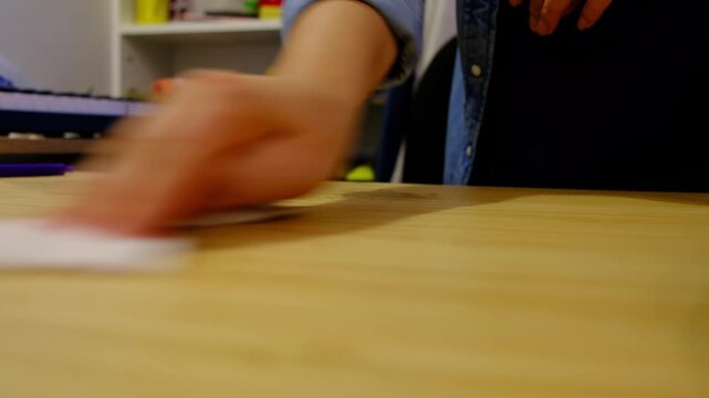 Low angle closeup shows a hand wiping a wooden table with a cloth, shallow focus and a calm indoor background. Regular cleaning reduces germs, supports hygiene, and keeps workspaces stress free.