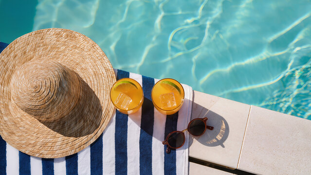Two orange cocktails with ice beside straw hat and sunglasses by pool. Summer resort flat lay. Tropical vacation refreshment. Poolside travel