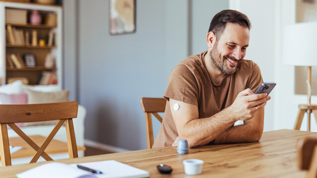 Smiling Man With Diabetes Using Smartphone and Continuous Glucose Monitor at Home Table