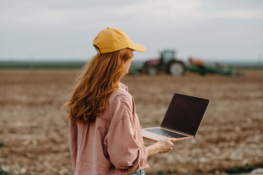 Young woman holding laptop in cultivated field near tractor