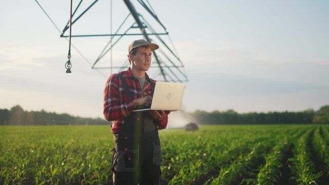 corn agriculture. a male farmer works on a laptop in a field with green corn sprouts. corn is watered by irrigation machine. irrigation agriculture sunlight business concept