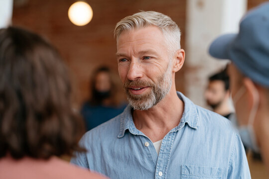 Man with graying hair and beard smiles warmly. He wears a light blue button-down shirt. He converses with two people in a casual setting. Soft lighting highlights his friendly expression