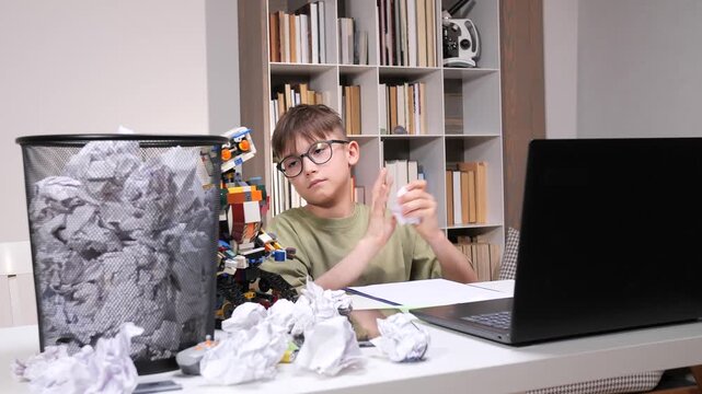 Young boy with glasses at a cluttered desk, laptop open, crumpling paper in frustration amid piles of failed drafts and a full waste bin during stressful home studying session