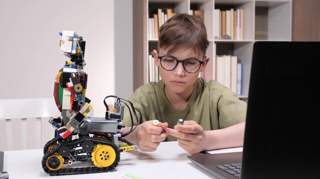 Young boy wearing glasses engaging in stem education, building a colorful robot, and holding parts while looking at a laptop, demonstrating hands on learning and technological development