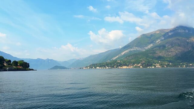 Panoramic scenery of Lake Como with high mountains touching clouds and colorful towns along the green coastline in Italy.