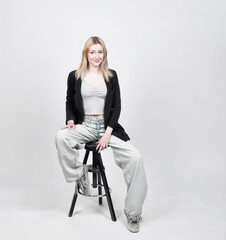 Fototapeta premium Junge blonde Frau sitzt entspannt auf Hocker vor hellem Studiohintergrund – modernes Lifestyle- und Fashion-Porträt mit natürlicher Ausstrahlung und viel copyspace und copy space.