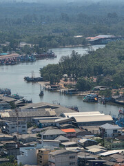 Fototapeta premium Aerial view showing calm peaceful coastal town with flowing river passing through industrial warehouse building and residential house near dense green forest under hazy daytime sky