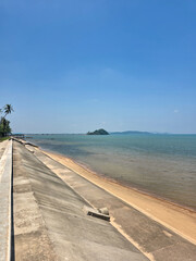 Fototapeta premium Beautiful serene beach scene featuring calm ocean water gently washing onto sandy shore beside sloping concrete sea wall under clear bright blue sky creating peaceful relaxing tranquil atmosphere