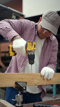 Confident carpenter man using cordless power drill to make hole in laminated wood board inside timber workshop. DIY woodworking, handmade furniture, home improvement and craftsmanship concept.