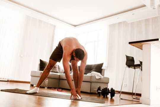 Young man go in for sport at home. Stand on mat and stretch down to toes. T-shirtless guy sportsman warming out body before exercise. Working out alone in middle of room. Sport ewuipment on floor.