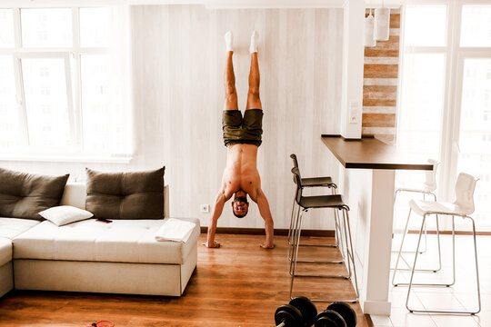 Athletic bodybuilding young man stands in his arms at home. Male sports model has a workout in his apartment in the living room. Young ordinary guy go in for sport at home
