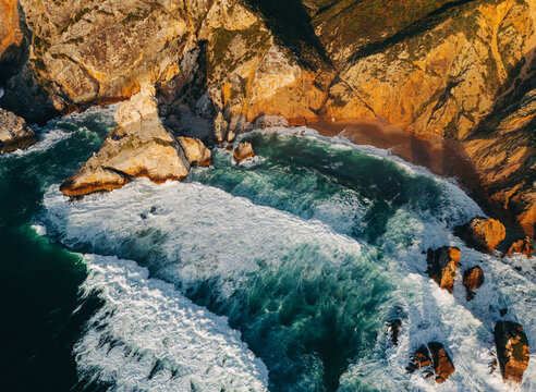Aerial view of dramatic rocky coastline with crashing ocean waves and golden sunset light over rugged sea cliffs