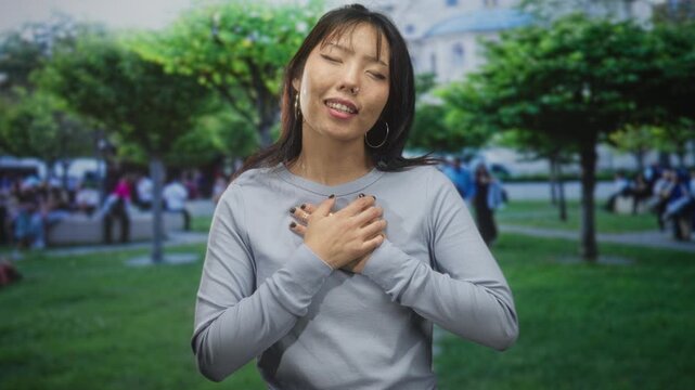 Young asian woman holds hands to chest with closed eyes and septum ring, standing in front of a building with trees and people nearby; gratitude mindfulness.