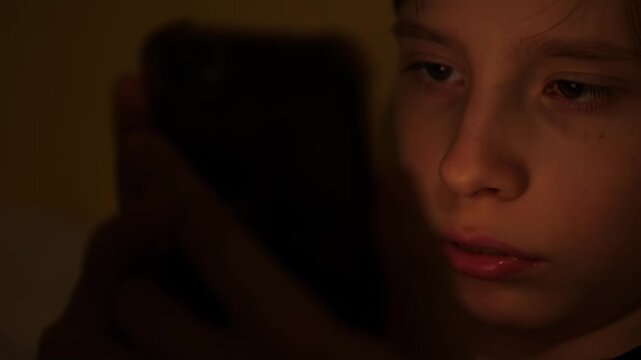 Close-up portrait of a child focused on a smartphone screen in complete darkness. Teenager using a mobile device at night illuminated by the dim light of the display