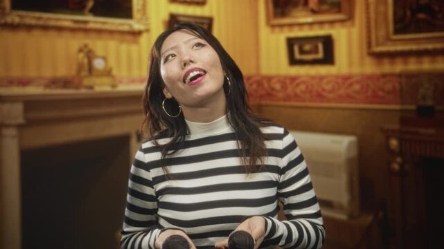 Young chinese woman in black and white striped top holding two chocolate muffins, tilting head back with mouth open and raising one cupcake toward her lips in ornate building; playful.