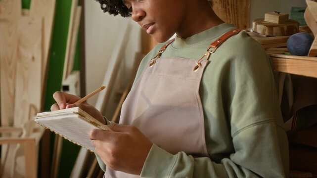 Focused African American teenage boy wearing apron writing in notebook with pencil at lesson in woodworking school