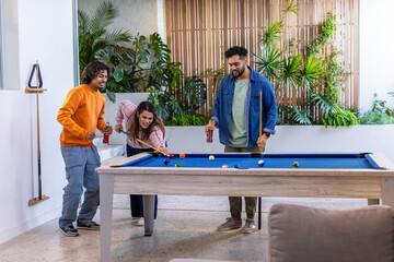 Naklejka premium Diverse friends leaning, taking shots and chatting in lounge with blue-felt pool table and cues