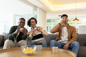Three adult males sitting on gray sofa in bright living room holding white controllers eating chips © wavebreak3