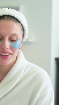 Vertical video: Pressing hand placing blue eye pads onto woman in robe, headband at home for care