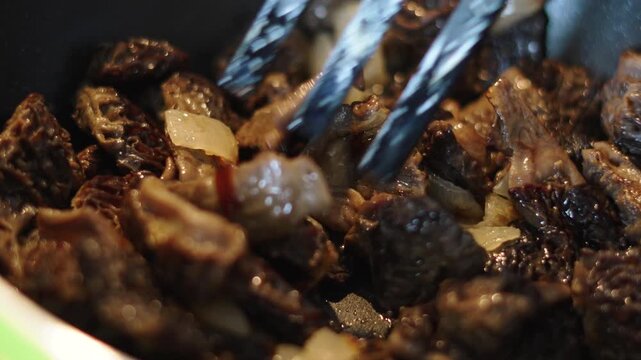 Stirring forest mushrooms with fork spatula during frying with onion, closeup. Cooking delicate morels in pan. Frying american forest mushrooms in oil, cooking high protein and high calorie food.