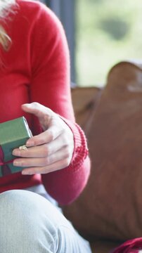 Vertical video: Seeing tear, woman in red top rotating and lifting green gift on sofa to inspect