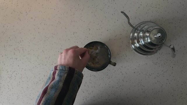 Unrecognizable Man in Striped Robe Stirring Hot Black Tea with Metal Spoon then Taking Mug Away from White Kitchen Table Top Down View POV High Angle Shot Finishing Morning Domestic Routine Shot
