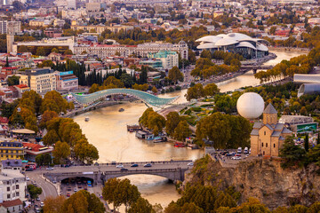 A wide view of Tbilisi, Georgia, shows Mtkvari (Kura) river flowing through the city, Metekhi church on a cliff, and Bridge of Peace. Various buildings and trees line the banks.  © Sergo