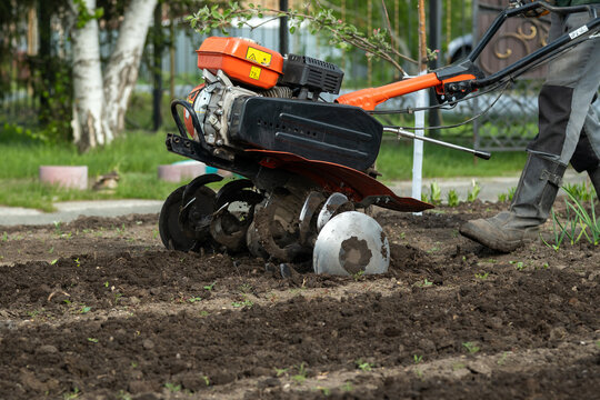 Gardening worker tilling ground with power tiller in spring season. A medium shot showing the lower half of a gardener in boots operating a powerful red tiller. 
