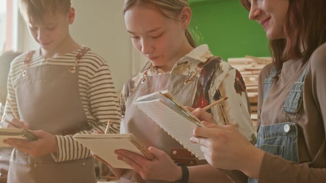 Three teenagers holding notebooks drawing sketches with pencils during woodworking masterclass