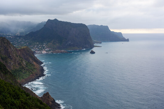 Vereda do Larano Trail overlooking Madeira's rugged coastline. Steep cliffs meet the Atlantic Ocean. Lush greenery frames the scenic mountain path. Cloudy skies above the dramatic seascape.