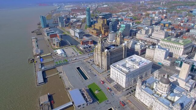 Breathtaking 4K aerial drone perspective of the historic Liverpool waterfront featuring the Three Graces at Pier Head under a clear blue sky, showcasing classic British architecture and city.