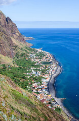 Fototapeta premium Madeira coastal village. White houses cluster along a curved black pebble shoreline on northern Madeira. Steep cliffs rise behind the settlement with terraced green slopes.
