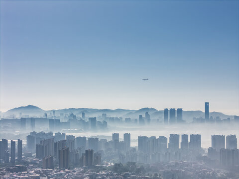 Aerial View of Modern Cityscape with High-Rise Buildings Emerging from Morning Fog and a Distant Airplane in the Sky