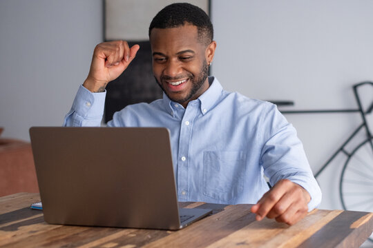 A smiling young man celebrates a moment of success while working on a laptop in a casual home office, conveying remote work productivity, online communication, confidence and positive professional ach