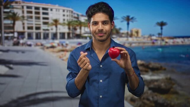 Young hispanic man holding red heart and making a finger heart gesture on a sunny street by the beach; affection connection.