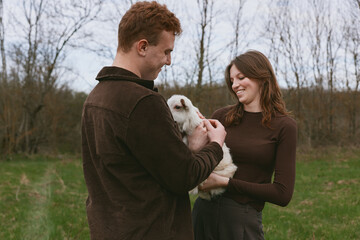 Fototapeta premium Couple outdoors with goat kid, sharing a gentle moment in a grassy field, capturing a warm lifestyle scene with a candid, natural feel and genuine connection between people and animal companions.