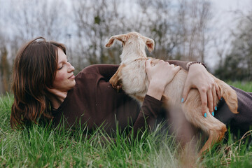 Fototapeta premium Woman lying on grass, cuddling a young dog in a tender moment. Outdoor setting conveys relaxation, affection, and simple lifestyle. Cozy, intimate scene with soft light and natural textures.