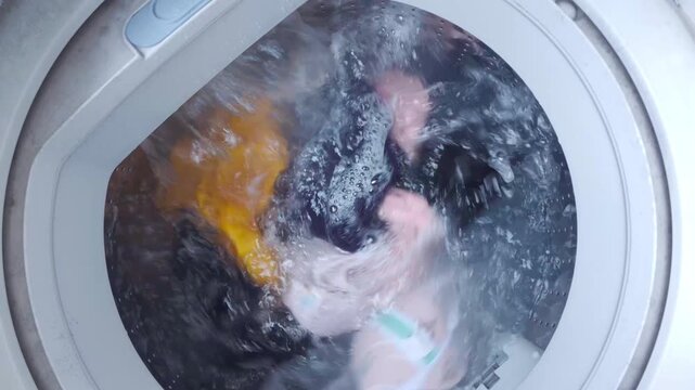 Top view of colorful clothes and soapy water spinning inside a top-loading washing machine drum during wash cycle.