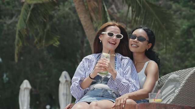 Beautiful diverse young lesbian couple holding drinks, smiling, and looking at something away in the distance while relaxing together on a beach lounge chair at a tropical resort during their romantic