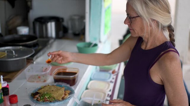 Mature woman prepares her shrimp tacos with various sauces at a taco stand in Baja California, Mexico during a lunchtime visit.