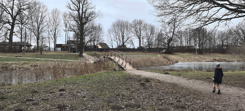 Child walks alone near a small wooden bridge by a pond on a cloudy day in a rural area with bare trees and grass
