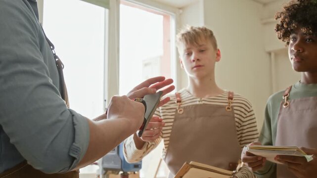 Two multiethnic teenage boys wearing aprons listening to unrecognisable male teacher describing parts of handplane at woodworking lesson